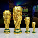 Set of gold World Cup trophies on a table with a sports event in the background.